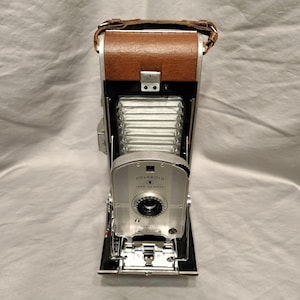 May include: A vintage Polaroid Land Camera Model 80 featuring a silver metal front plate with black text, a brown textured leather top cover, a brown leather carrying strap, and a silver accordion-style bellows mechanism extending from the body.