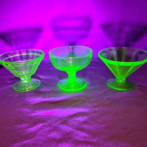 May include: Three glass dessert bowls glowing with a bright neon green color under ultraviolet light. Each bowl features a distinct shape, including one with vertical ribbing, one with a smooth rounded basin, and one with a wide, angled rim.