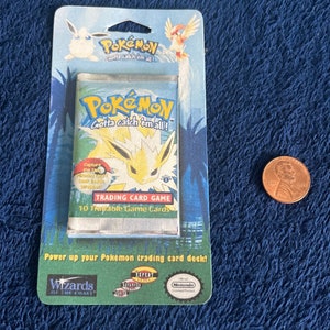 Puede incluir: Un paquete sellado de cartas de juego de cartas coleccionables Pokémon con un personaje Pokémon amarillo y blanco en la parte delantera. El paquete está etiquetado como "Gotta catch 'em all!" e incluye 10 cartas de juego intercambiables. El paquete está sobre un fondo verde y azul con el logotipo de Wizards of the Coast y el logotipo de Nintendo.