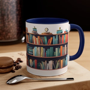 Puede incluir: Taza de cerámica blanca con borde y asa azul marino. La taza presenta una ilustración colorida de una estantería llena de libros.