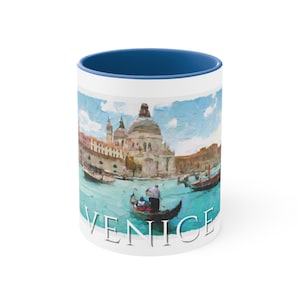 May include: White ceramic mug with a blue rim. The mug features a watercolor painting of a canal in Venice, Italy. The painting includes a gondola with a gondolier, a church, and buildings. The text "VENICE" is printed on the bottom of the mug.