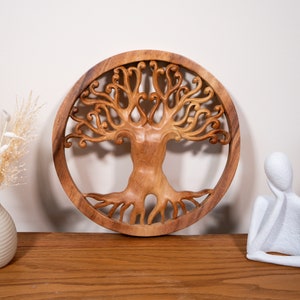 Può includere: Un'appendice da parete rotonda in legno con un design a forma di albero della vita intagliato. L'albero ha rami e radici intricati, e il legno è di un colore marrone caldo.