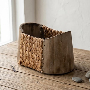 Puede incluir: Una cesta de madera con un panel tejido, de color marrón claro y texturizado en la parte delantera. La cesta tiene una parte superior curvada y está colocada sobre una superficie de madera. La cesta es de color natural y neutro.