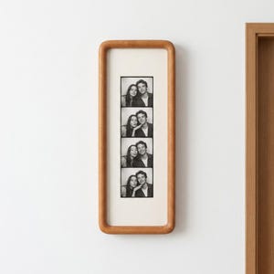 Op de afbeelding: Een verticale rechthoekige houten fotolijst met afgeronde hoeken met daarin een zwart-wit fotostrip. De strip bevat vier gestapelde portretten van een man en een vrouw die samen poseren met verschillende uitdrukkingen tegen een witte passe-partout.
