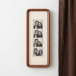 Könnte beinhalten: Ein vertikaler, rechteckiger Bilderrahmen aus Holz mit abgerundeten Ecken und einem cremefarbenen Passepartout. Im Rahmen befindet sich ein schwarz-weißer Fotostreifen mit vier übereinander angeordneten Porträts eines Paares mit verschiedenen Gesichtsausdrücken.