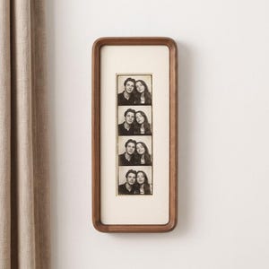 Könnte beinhalten: Ein vertikaler, rechteckiger Bilderrahmen aus Holz mit abgerundeten Ecken und einem hellen Passepartout. Im Rahmen befindet sich ein schwarz-weißer Fotostreifen mit vier übereinander angeordneten Porträts eines Mannes und einer Frau.