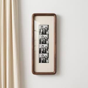 May include: A vertical rectangular wooden picture frame with rounded corners, featuring a light-coloured mat board. Inside the frame is a black and white photo strip containing four stacked images of three people posing together in various expressions.