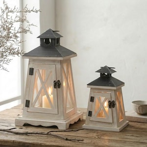 May include: Two square lanterns with distressed white wooden frames and dark metal roofs. Each lantern features a hinged door with a metal latch, X-shaped crossbars on the sides, and a single lit pillar candle inside the glass-paneled enclosure.