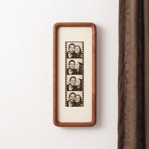 Puede incluir: Un marco de madera rectangular vertical con esquinas redondeadas que contiene una tira de fotos en blanco y negro de fotomatón. La tira presenta cuatro cuadros apilados que muestran a una pareja posando junta con diversas expresiones sobre un fondo crema.