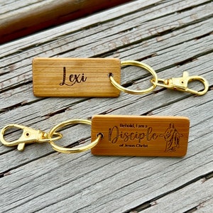 May include: Two wooden keychains with gold rings and clasps. The first keychain has the name "Lexi" engraved on it. The second keychain has the text "Behold, I am a Disciple of Jesus Christ" engraved on it, along with a small image of Jesus.