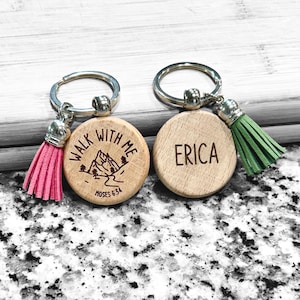 May include: Two wooden keychains with silver keyrings and tassels. One keychain has a pink tassel and the text "WALK WITH ME" with a mountain graphic. The other keychain has a green tassel and the name "ERICA".
