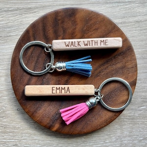 May include: Two keychains with wooden rectangular tags and silver keyrings. One keychain has the text "WALK WITH ME" and a blue tassel. The other has the name "EMMA" and a pink tassel. Both are on a wooden surface.
