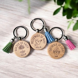 May include: Three wooden keychains with engraved inspirational phrases and matching tassels. The keychains feature phrases like "Builder of Faith," "Messenger of Hope," and "Gatherer of Light." Each keychain has a different colored tassel: green, blue, and pink.