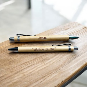 May include: Two bamboo pens with silver accents and black ink tips. One pen is engraved with the name "Jessa", and the other with the phrase "Walk with me". The pens are lying on a wooden surface.