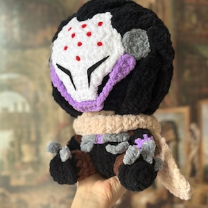 May include: A handmade crochet doll featuring a black head with a white mask, red dots, and purple accents. The doll has a beige scarf and brown and gray details. The doll is held in a person's hand.