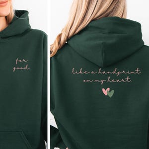 Puede incluir: Sudadera con capucha verde bosque con bolsillo delantero y capucha. La parte delantera de la sudadera tiene el texto "for good" en cursiva. La parte trasera de la sudadera tiene el texto "like a handprint on my heart" con dos pequeños corazones.