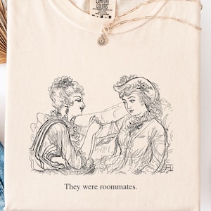 May include: Ivory-colored t-shirt with a black and white illustration of two women and the text "They were roommates." The shirt is laid flat with a pair of blue jeans, white shoes, and a necklace.
