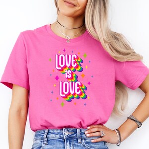 May include: A pink t-shirt with the text "LOVE IS LOVE" in white, rainbow, and star accents. The shirt is short-sleeved and is paired with blue jeans. The words "Charity Pink" are written in cursive on the top left.