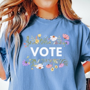 Op de afbeelding: Een lichtblauw T-shirt met het woord "VOTE" in het wit, omringd door een bloemenkrans van kleurrijke bloemen en groene bladeren. Het shirt heeft een casual stijl.