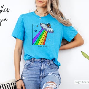 Pu&ograve; includere: Maglietta Heather Aqua turchese con una grafica colorata di un UFO che emette un raggio arcobaleno. Il design include pianeti e la scritta "Happy Pride".