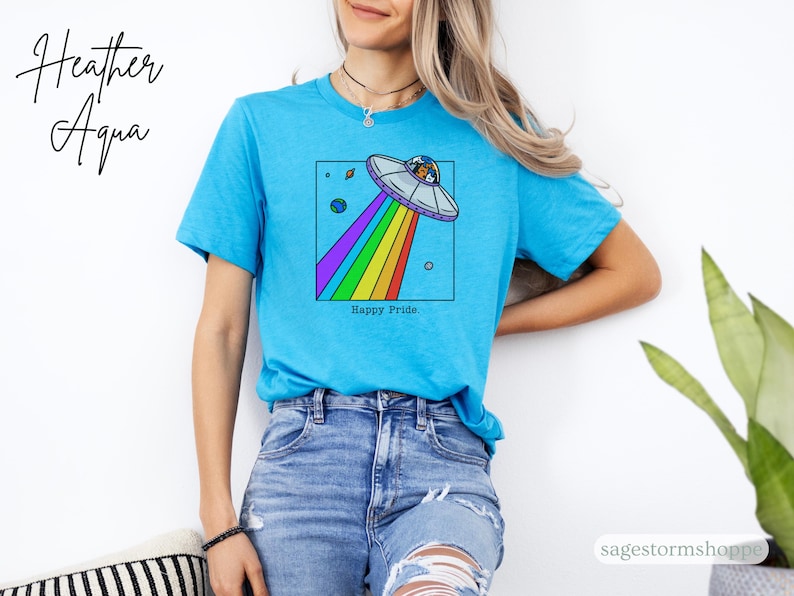 Pu&ograve; includere: Maglietta Heather Aqua turchese con una grafica colorata di un UFO che emette un raggio arcobaleno. Il design include pianeti e la scritta "Happy Pride".