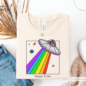 Pu&ograve; includere: T-shirt color crema con una grafica di un UFO che emette un arcobaleno. Il design include pianeti e la scritta "Happy Pride". La maglietta &egrave; esposta con jeans blu e scarpe da ginnastica bianche.