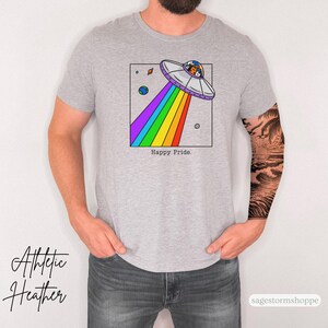 Pu&ograve; includere: T-shirt grigio athletic heather con una grafica colorata di un UFO che emette un arcobaleno. Il design include pianeti e la scritta "Happy Pride". La maglietta &egrave; un capo d'abbigliamento casual.
