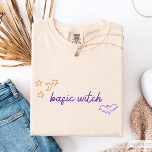 Peut inclure: T-shirt couleur ivoire avec l'inscription "basic witch" en violet, accompagnée d'étoiles orange et d'un croissant de lune. Un motif de chauve-souris violet est également présent. Le t-shirt est plié, à côté d'un jean, de chaussures blanches et d'un collier.