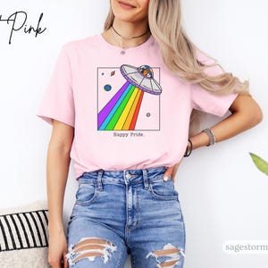 Pu&ograve; includere: T-shirt rosa chiaro con una grafica di un UFO che emette un raggio arcobaleno, con la scritta "Happy Pride". La persona indossa jeans blu.