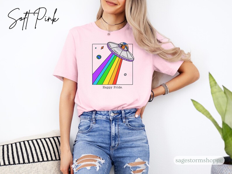 Pu&ograve; includere: T-shirt rosa chiaro con una grafica di un UFO che emette un raggio arcobaleno, con la scritta "Happy Pride". La persona indossa jeans blu.