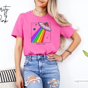 Pu&ograve; includere: Una maglietta rosa con una grafica di un UFO che emette un raggio arcobaleno. Il testo "Happy Pride" &egrave; sotto la grafica. La scritta "Charity Pink" &egrave; scritta in corsivo in alto a sinistra.