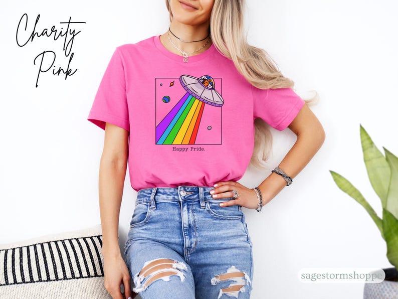 Pu&ograve; includere: Una maglietta rosa con una grafica di un UFO che emette un raggio arcobaleno. Il testo "Happy Pride" &egrave; sotto la grafica. La scritta "Charity Pink" &egrave; scritta in corsivo in alto a sinistra.