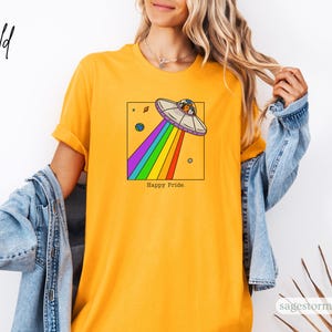 Pu&ograve; includere: T-shirt giallo senape con un design a forma di UFO a cartoni animati, che emette un arcobaleno, e la scritta "Happy Pride". L'UFO &egrave; all'interno di un quadrato nero, con pianeti sullo sfondo. Un capo casual.