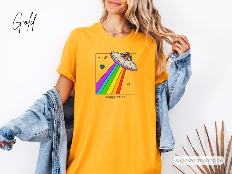 Pu&ograve; includere: T-shirt giallo senape con un design a forma di UFO a cartoni animati, che emette un arcobaleno, e la scritta "Happy Pride". L'UFO &egrave; all'interno di un quadrato nero, con pianeti sullo sfondo. Un capo casual.