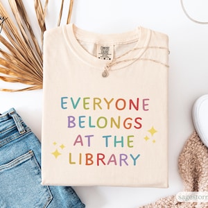 Op de afbeelding: Ivoor kleurig t-shirt met de tekst "EVERYONE BELONGS AT THE LIBRARY" in regenboogkleuren. Het shirt wordt gecombineerd met een blauwe jeans, witte sneakers en een ketting. Het ontwerp bevat twee gele sterren.