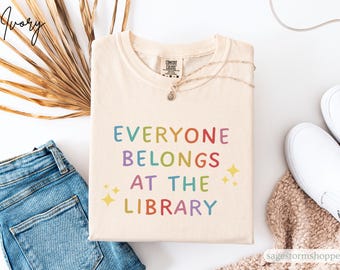 Puede incluir: Camiseta color marfil con el texto "EVERYONE BELONGS AT THE LIBRARY" en colores arcoíris. La camiseta se combina con vaqueros azules, zapatillas blancas y un collar. El diseño incluye dos estrellas amarillas.