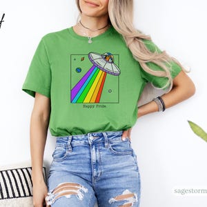 Pu&ograve; includere: T-shirt verde con un'illustrazione a cartoni animati di un UFO con un raggio arcobaleno, all'interno di un quadrato nero. Il testo "Happy Pride" &egrave; in basso. La maglietta &egrave; di colore verde unito.