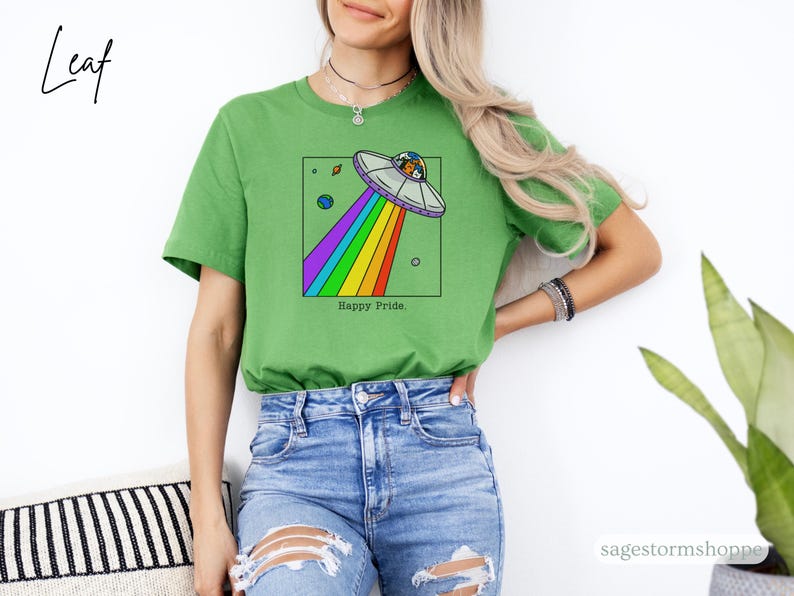 Pu&ograve; includere: T-shirt verde con un'illustrazione a cartoni animati di un UFO con un raggio arcobaleno, all'interno di un quadrato nero. Il testo "Happy Pride" &egrave; in basso. La maglietta &egrave; di colore verde unito.