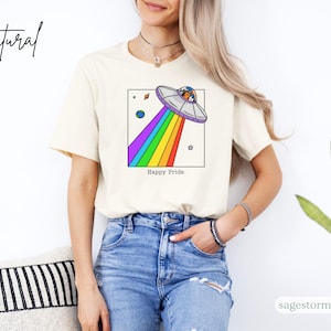 Pu&ograve; includere: T-shirt color crema con un'illustrazione a cartoni animati di un UFO con un raggio arcobaleno e le parole "Happy Pride". Il design include pianeti e stelle. La maglietta &egrave; indossata da una persona con jeans blu.