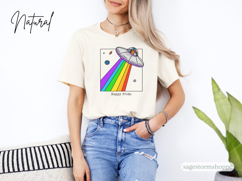 Pu&ograve; includere: T-shirt color crema con un'illustrazione a cartoni animati di un UFO con un raggio arcobaleno e le parole "Happy Pride". Il design include pianeti e stelle. La maglietta &egrave; indossata da una persona con jeans blu.