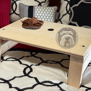 May include: A light-colored wooden lap desk with four sturdy legs and a smooth rectangular surface. The top features a circular cutout and a circular logo with a dog illustration and the text WoodDog Crafts. A pair of brown sandals rests on top.
