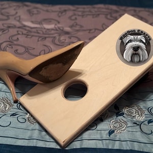 May include: A light-colored wooden cornhole board with a round hole and a high-heeled shoe. The board has a circular logo with a dog's face and the text "Woodog Crafts". The shoe is beige.
