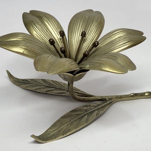 Puede incluir: Una escultura de flor de metal color latón, con seis pétalos y dos hojas. La flor tiene un diseño detallado con vetas visibles en los pétalos y las hojas. La escultura es probablemente un artículo decorativo.