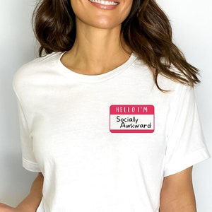 May include: White t-shirt with a pink name tag that reads "HELLO I'M Socially Awkward". The shirt is paired with blue jeans and a red, black, and white plaid shirt tied around the waist.
