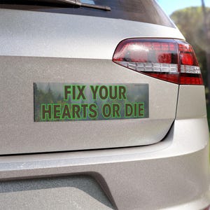 Pode incluir: Um carro prateado com um adesivo de pára-choques que diz "Fix Your Hearts Or Die" em texto marrom e verde sobre um fundo de floresta desfocado.