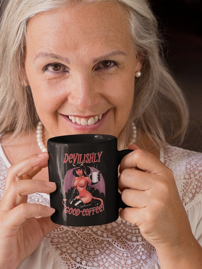 May include: Black coffee mug with a white design of a woman with red horns and a tail holding a coffee cup. The text on the mug reads "Devilishly good coffee!"