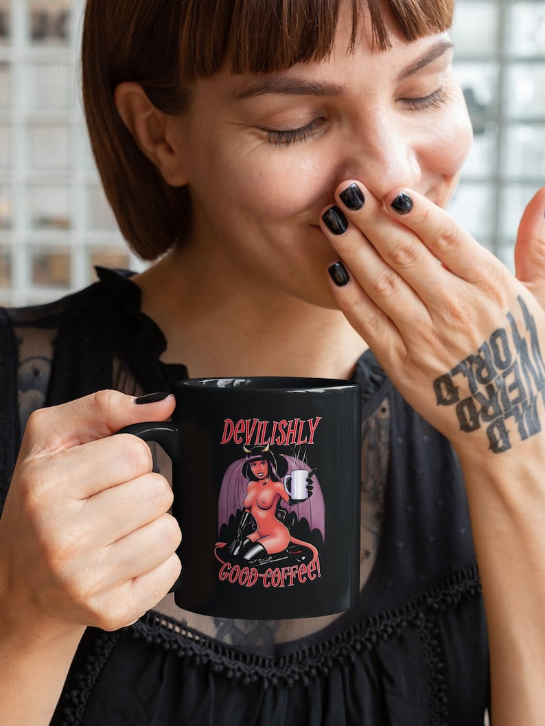 May include: Black coffee mug with a red and black illustration of a woman with devil horns and wings holding a coffee cup. The text on the mug reads "Devilishly good coffee!"