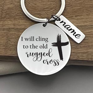 May include: A silver-colored keychain with a round pendant and a rectangular tag. The round pendant has the text "I will cling to the old rugged cross" with a black cross graphic. The rectangular tag has the word "name" in cursive.