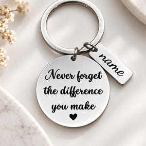 May include: A silver-toned metal keychain featuring a circular pendant engraved with the black text 'Never forget the difference you make' and a small heart symbol. Attached is a rectangular metal tag with the word 'name' engraved in black.