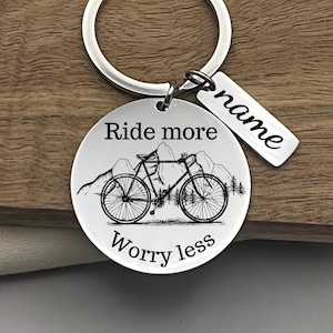 May include: A silver-colored keychain with a round pendant featuring a bicycle illustration, mountains, and the text "Ride more Worry less." A smaller rectangular tag is attached with the word "name" on it. The keychain is on a wooden surface.